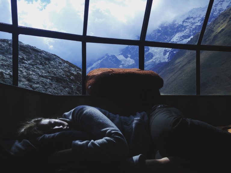 Woman sleeps under a glass ceiling. Outside, mountains and clouds are above her head.