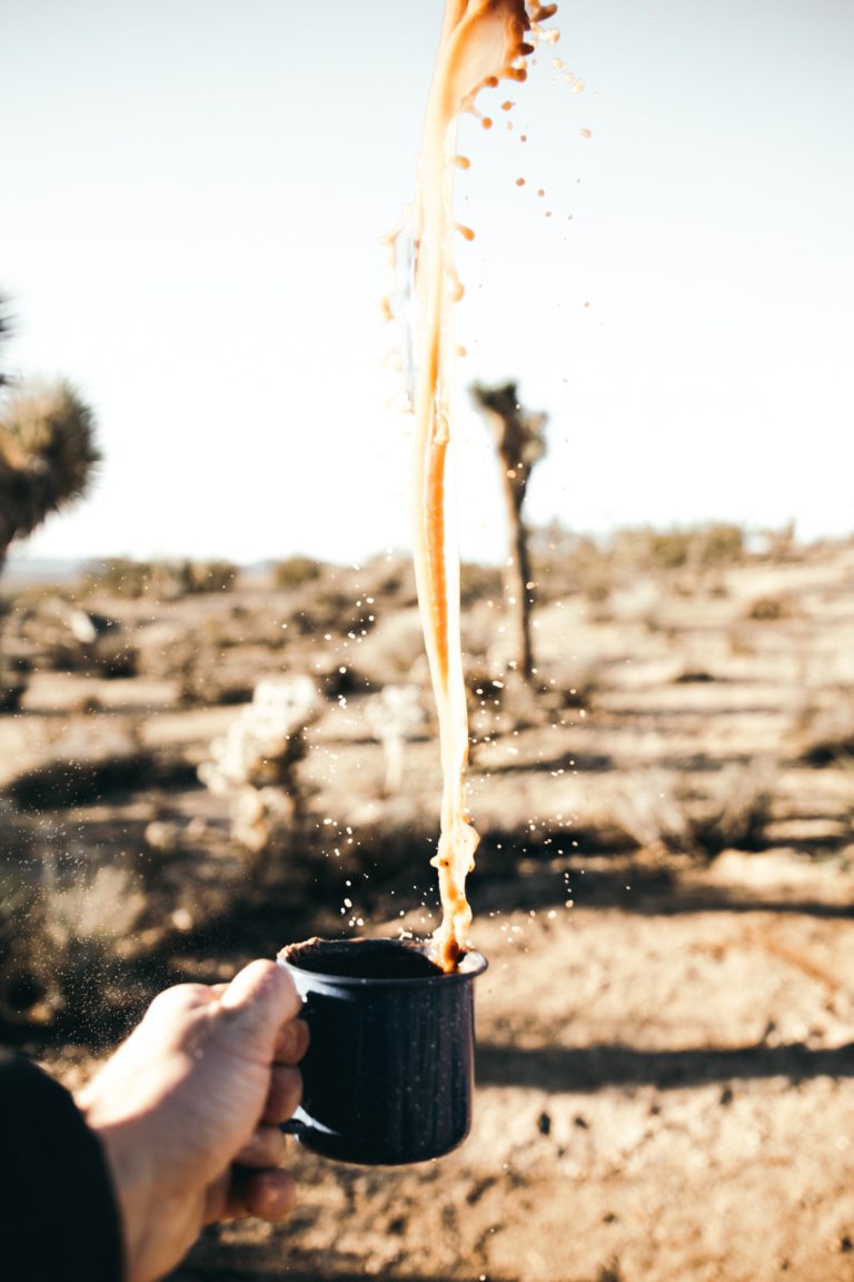 Hand holds a black mug that has coffee spilling out of it and up in the air. Faded desert landscape in the background.