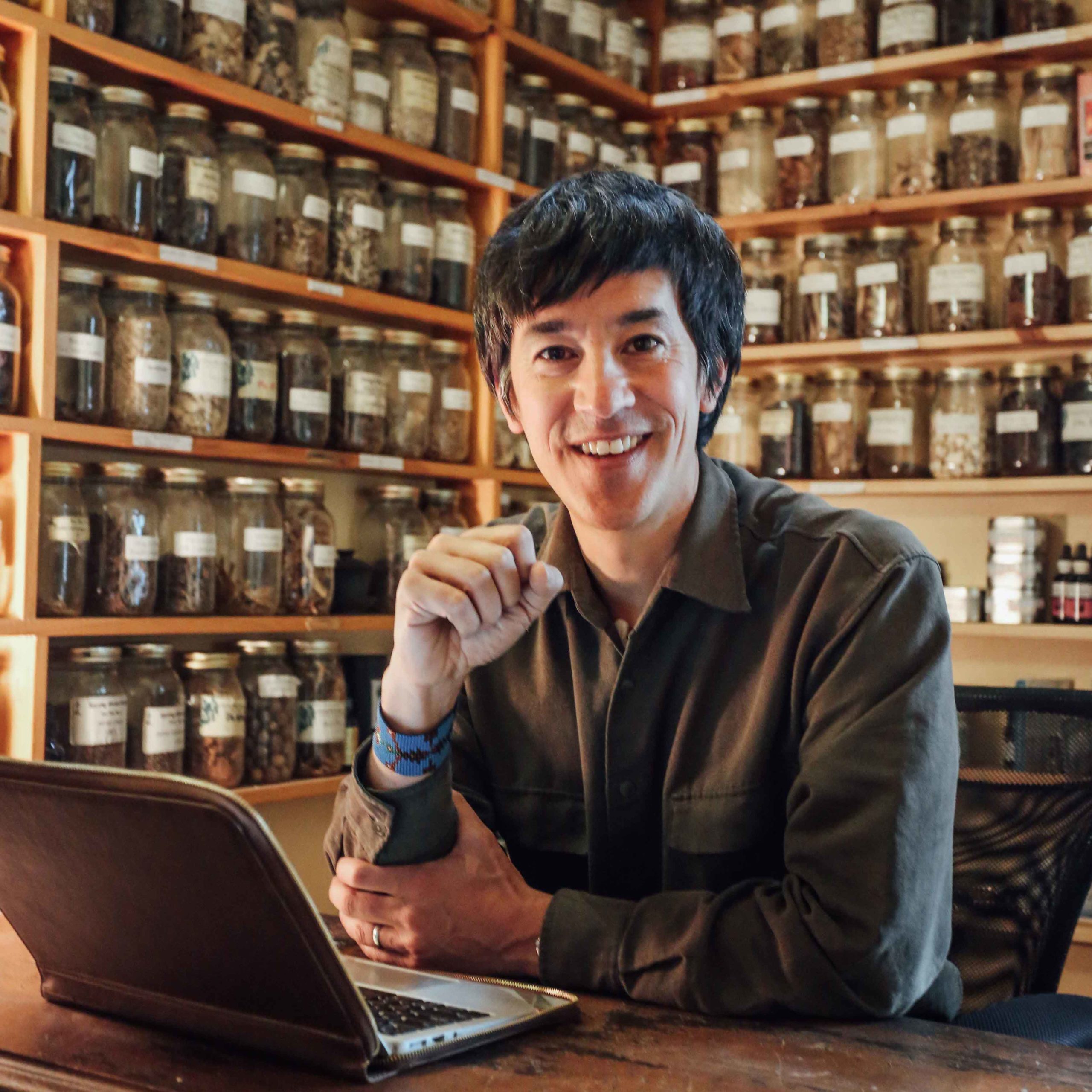Marco Lam sits at a desk, his computer is in front of him. In the background are shelves full of jars of Chinese herbs.